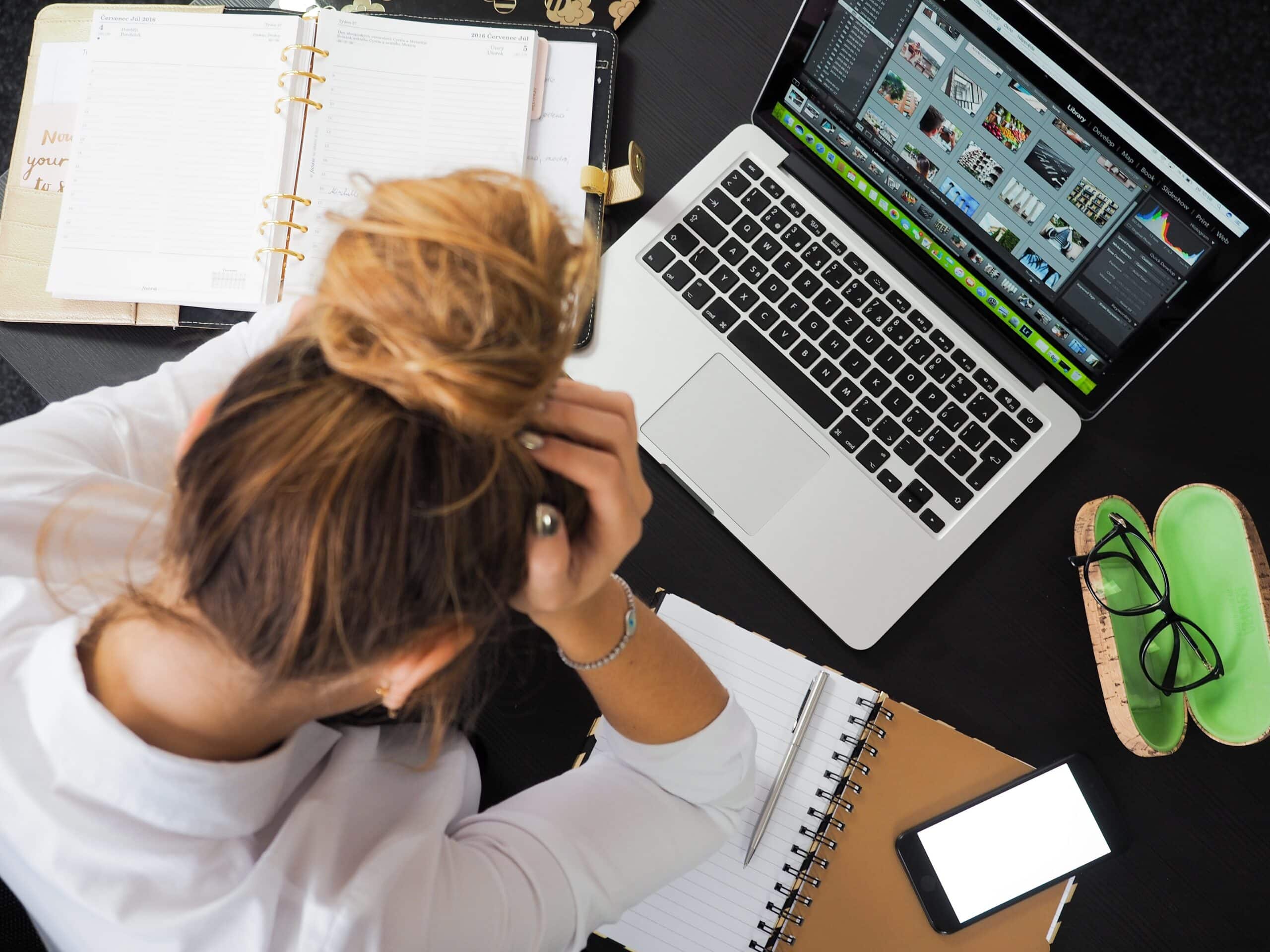 Eine erschöpfte Frau sitzt mit gesenktem Kopf an einem Schreibtisch vor ihrem Laptop.